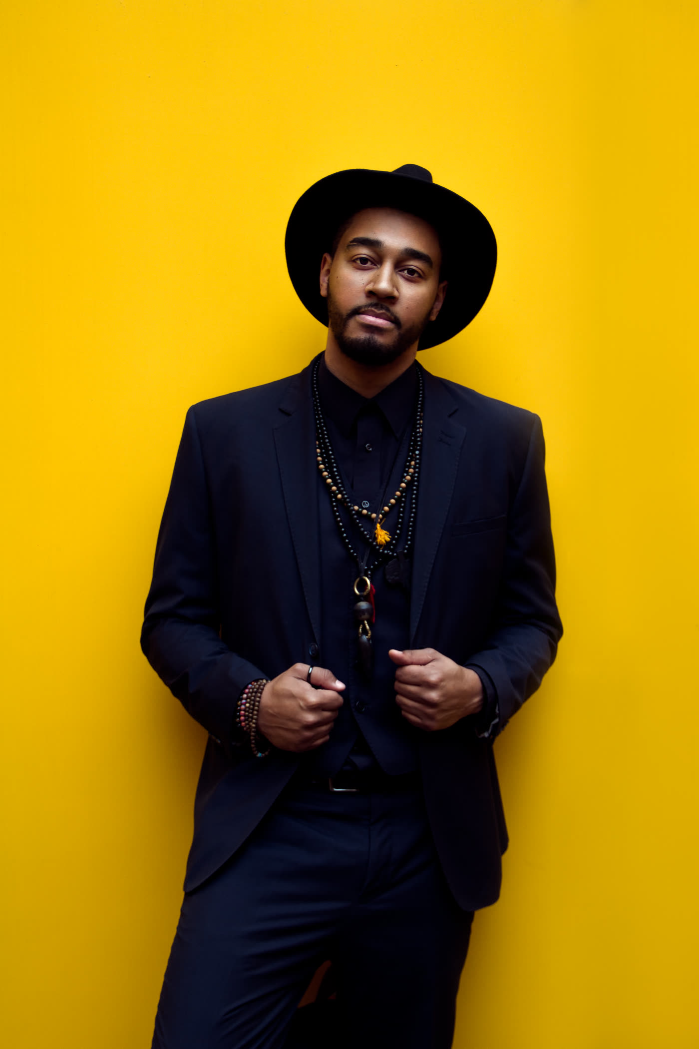 Kwame Aki, Melbourne wedding singer, in a dark suit and hat against a gold wall